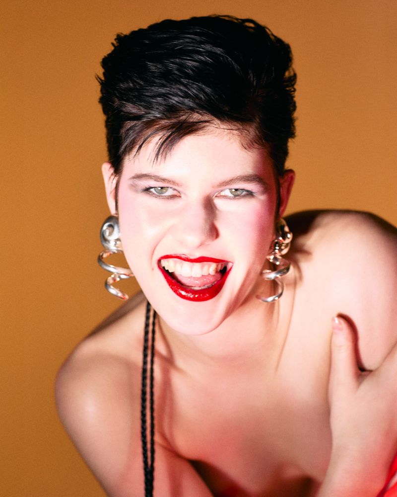 Headshot portrait of a smiling young adult female with short dark hair, large spiral earrings, and red lipstick against a gold background.