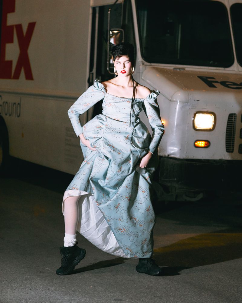 Woman in formal dress and sneakers posing in front of a FedEx truck with low lighting and night time urban setting.
