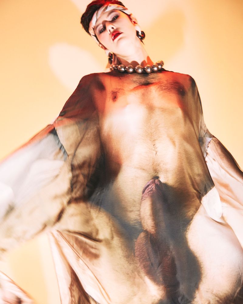 Model Rachel Goff wearing a translucent printed garment with large statement necklace and headband against a warm background.