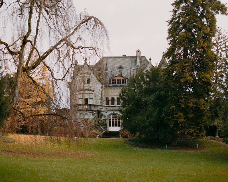 Elegant mansion with gray stone facade, multiple windows, and steeply pitched roof, surrounded by lush green lawn and mature trees under overcast sky.