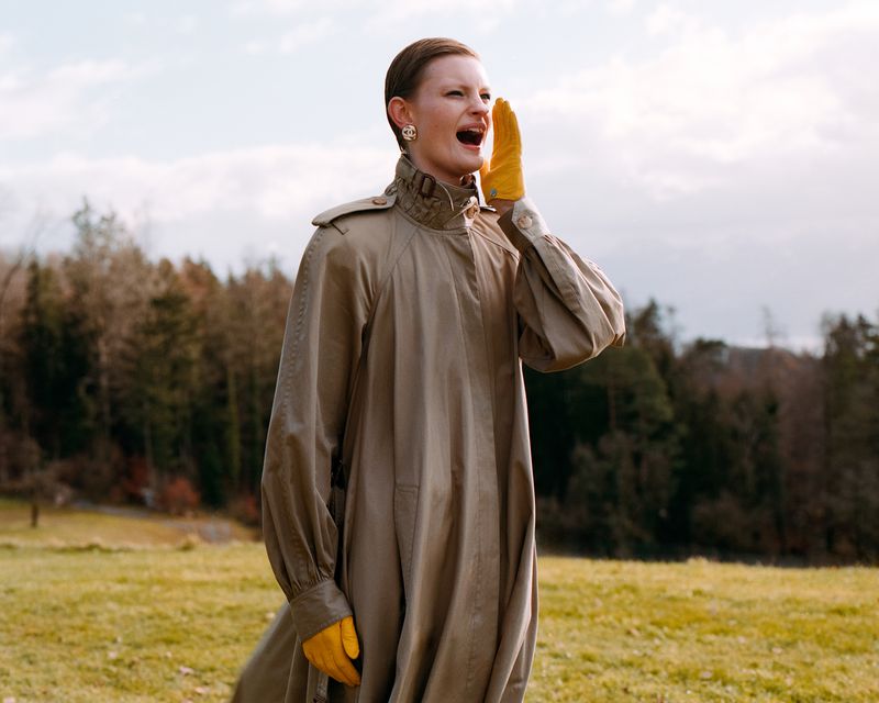 An adult female in a long beige trench coat yells outdoors in a field with trees in the background, wearing yellow gloves and earrings.