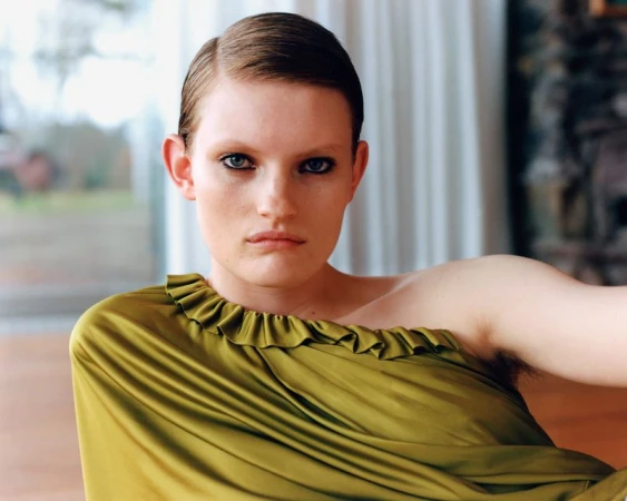 Elegant woman poses indoors wearing a green, one-shoulder gown. Soft, natural lighting and a blurred background create a sophisticated mood.
