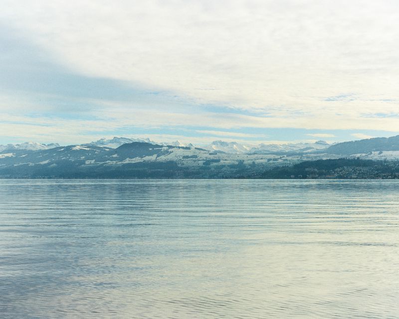 Scenic view of a tranquil lake under a cloudy sky with snow-capped mountains in the background, creating a serene and natural landscape.