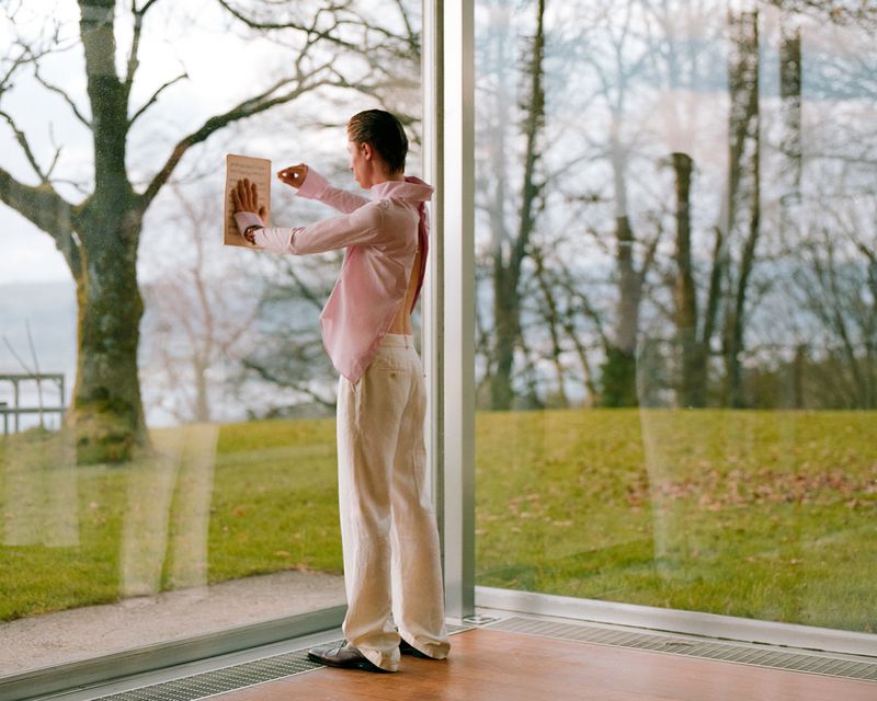 Model standing inside by a large window holding a sheet of music against the glass, overlooking a lawn and trees