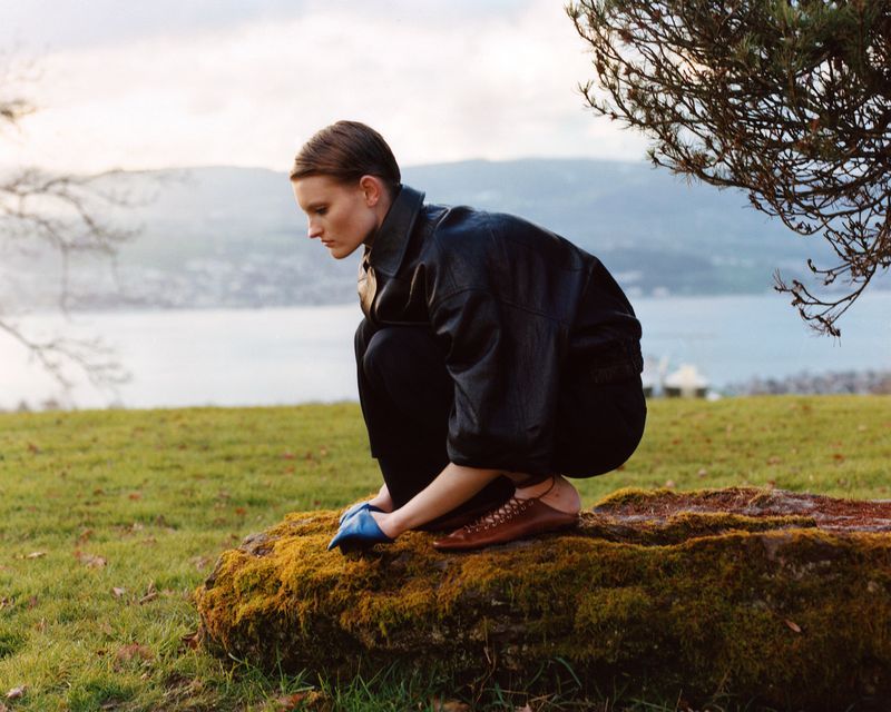 Woman in jacket crouches on a mossy rock outdoors with a lake and mountains in the background, dressed in black with tan shoes.