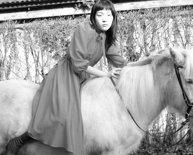 A monochromatic image shows a young Asian woman in a dress riding bareback on a white horse in front of a hedge outdoors.