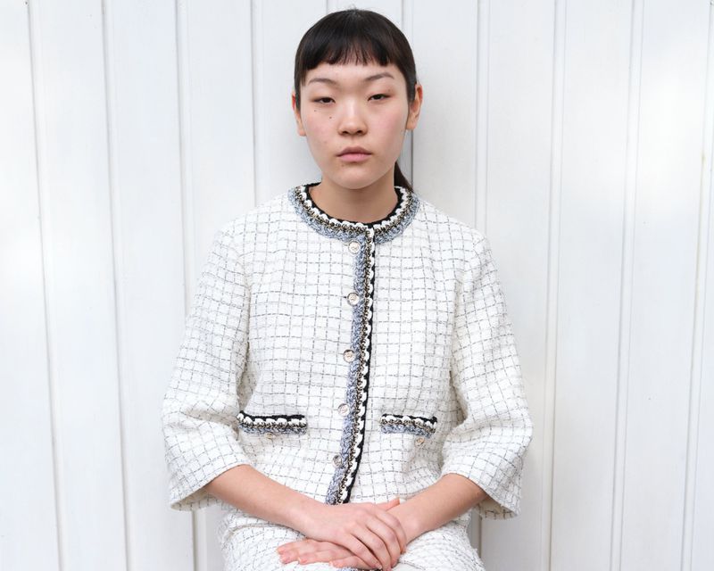 A young woman with dark hair is shown in a medium shot wearing a tweed jacket and skirt, posing against a white paneled background.