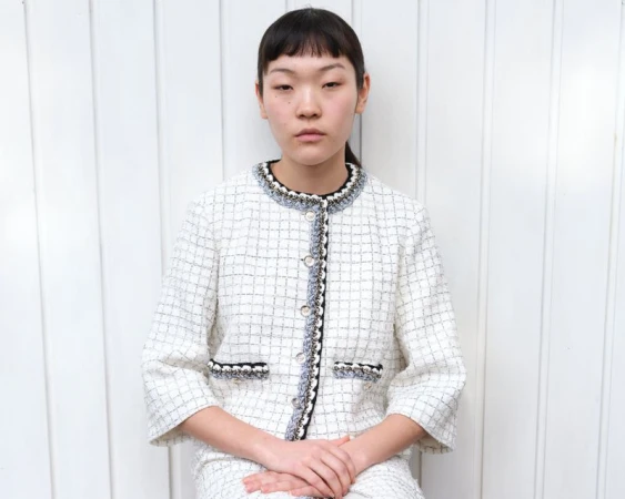 A young woman with dark hair is shown in a medium shot wearing a tweed jacket and skirt, posing against a white paneled background.