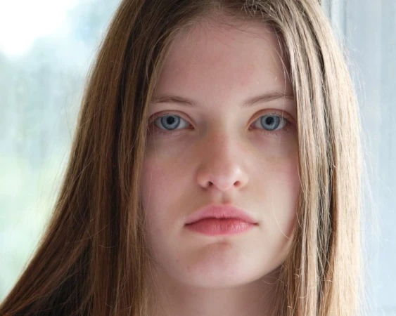 Close-up portrait of a young woman with long brown hair and blue eyes looking directly at the camera, set against a bright, blurred background.