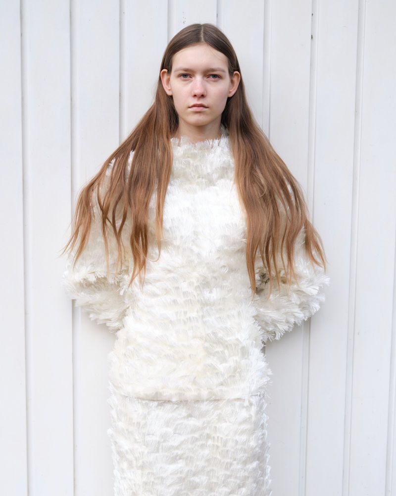 A young, fair-skinned woman with long brown hair stands in front of a white wooden wall, wearing a white two-piece feathery outfit.