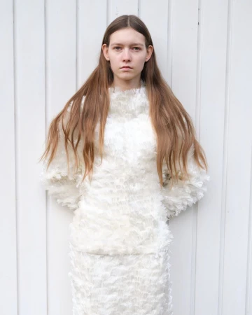 A young, fair-skinned woman with long brown hair stands in front of a white wooden wall, wearing a white two-piece feathery outfit.