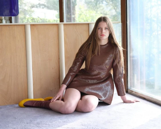 Young woman with long brown hair wearing a glossy brown dress and yellow boots seated on a carpeted floor indoors