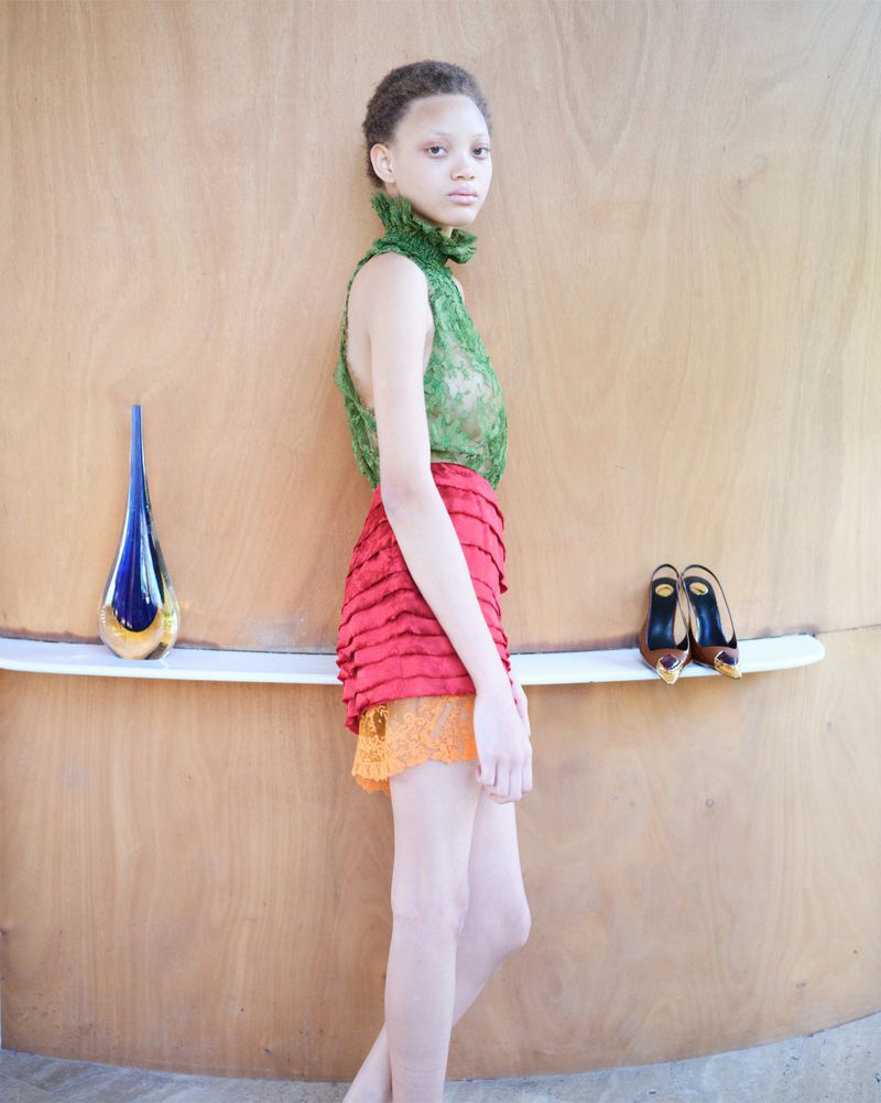 Young adult woman posing in green lace top and layered skirt in front of wood paneled wall with vase and heels on shelf.