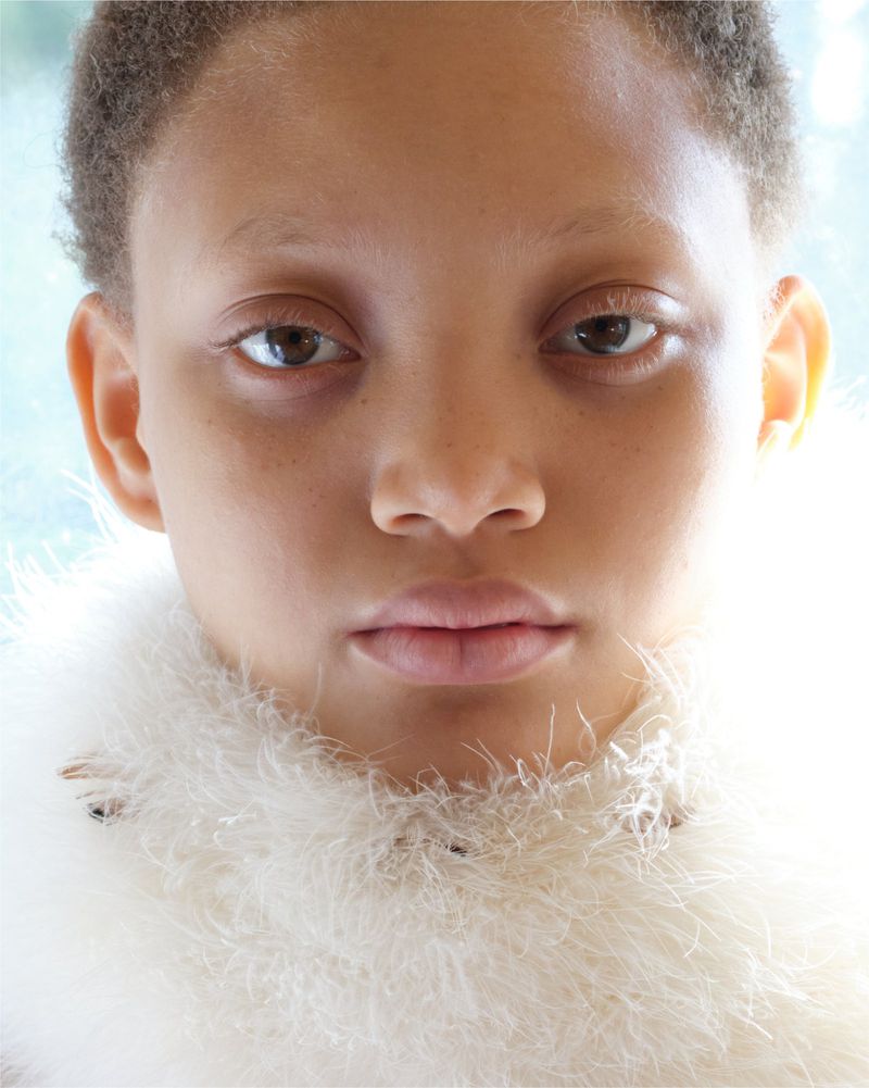 Close-up portrait of a young person with light skin, brown eyes, and short hair, wearing a feathery collar, set against a blurred background.