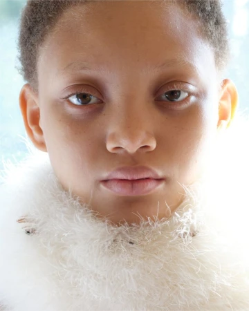 Close-up portrait of a young person with light skin, brown eyes, and short hair, wearing a feathery collar, set against a blurred background.