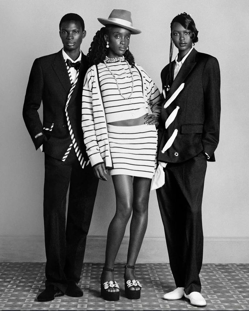 Three Black fashion models pose in a studio wearing black and white outfits, including a suit and a striped skirt and sweater set.