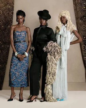 Three models wearing patterned blue dress, black suit with fringed stole, and light blue gown with fringed headpiece.