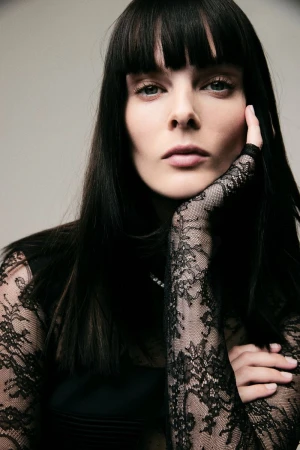 Model with dark hair and bangs resting her head on a hand wearing a black lace sleeve against a light grey background