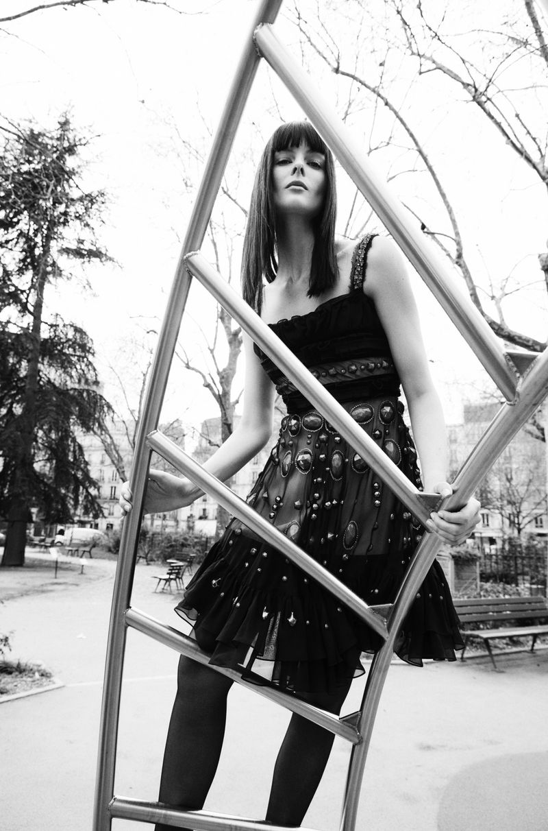 Monochrome image of a woman in a dark dress posing on metal playground equipment in a park setting with bare trees.