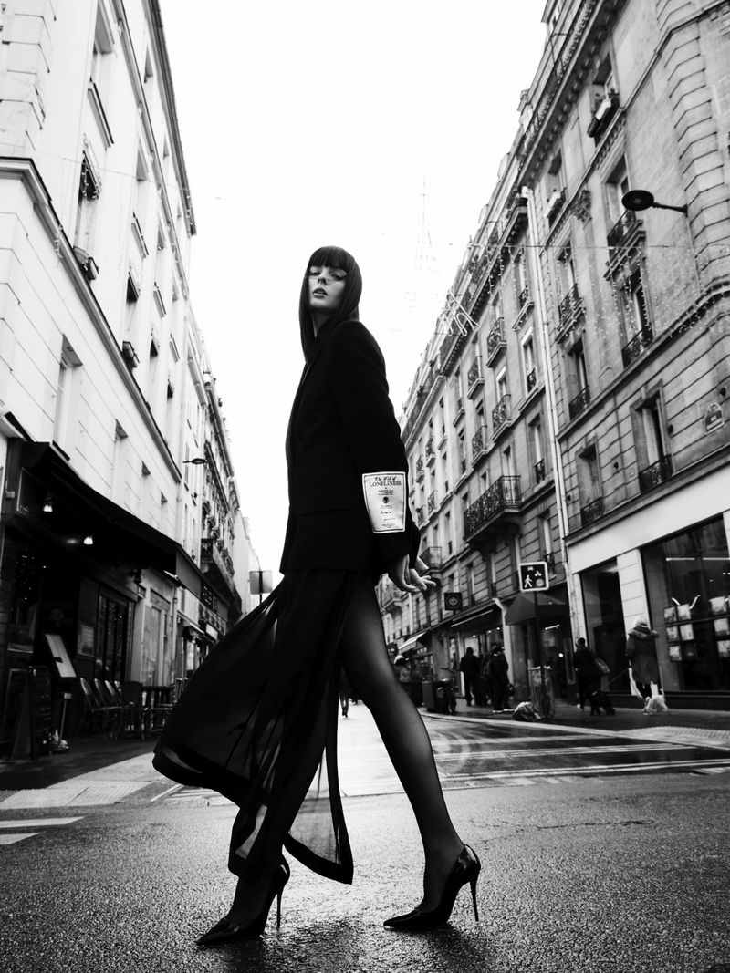 Fashion shot in black and white of a woman in a blazer and sheer skirt crossing a wet street between buildings in Paris, France.