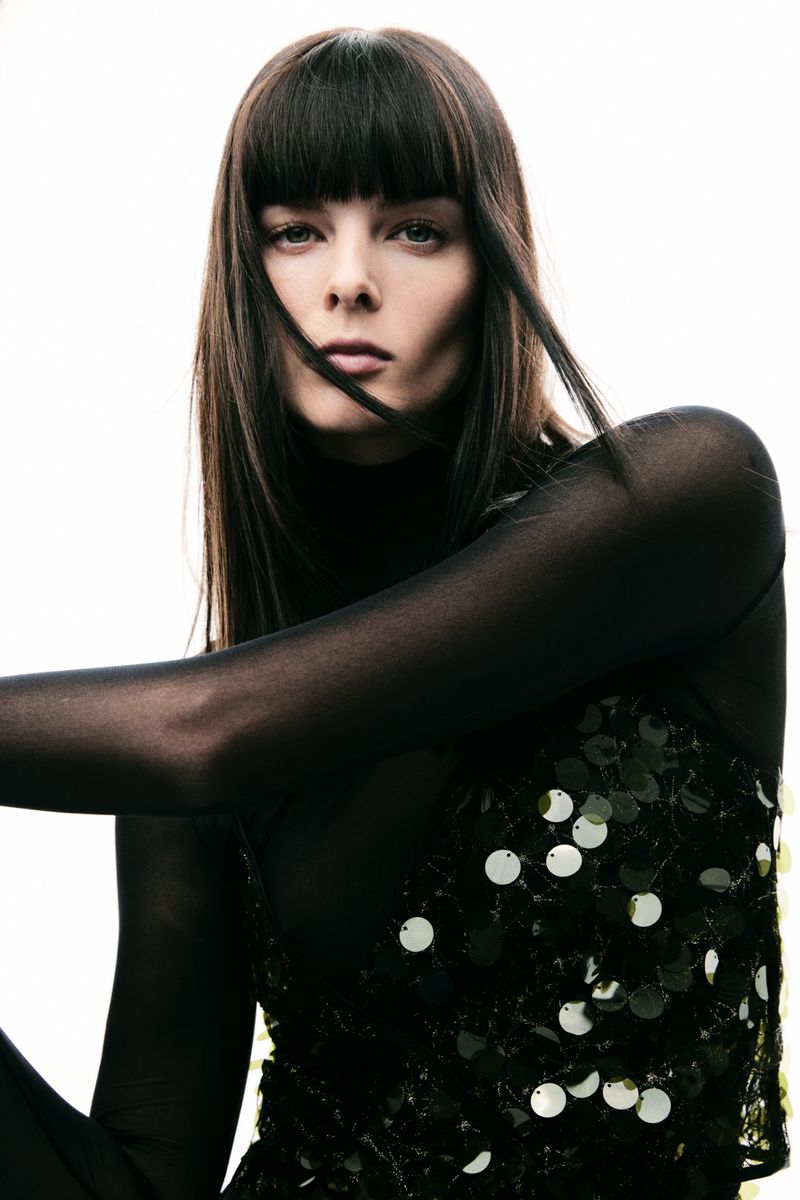 Portrait of a woman with long dark hair wearing a black mesh top with sequins against a bright white backdrop in a studio setting.