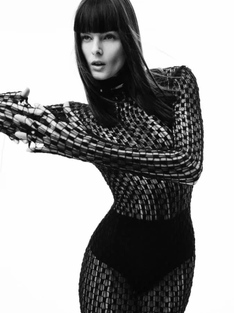 A fashion shot of a woman with dark hair wearing a long-sleeved, semi-sheer black dress against a bright white backdrop in a studio setting.