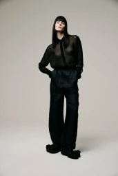 Model Aylah Peterson in a sheer black shirt and striped trousers posing against a grey studio seamless backdrop.