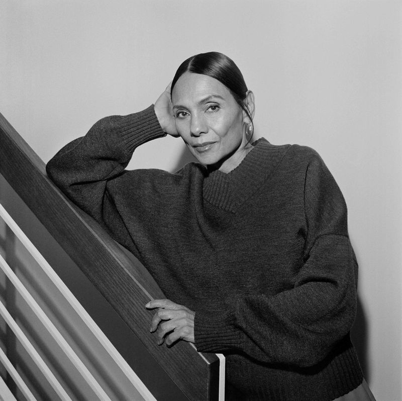 Portrait of a middle-aged woman with slicked-back hair, wearing an oversized sweater, leaning on a wooden staircase in a studio setting.