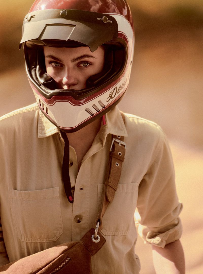 Close-up of a person wearing a helmet and a beige jumpsuit with a brown bag slung across the body, with a blurred background.