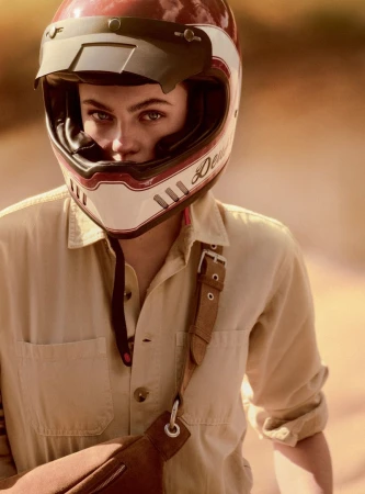 Close-up of a person wearing a helmet and a beige jumpsuit with a brown bag slung across the body, with a blurred background.