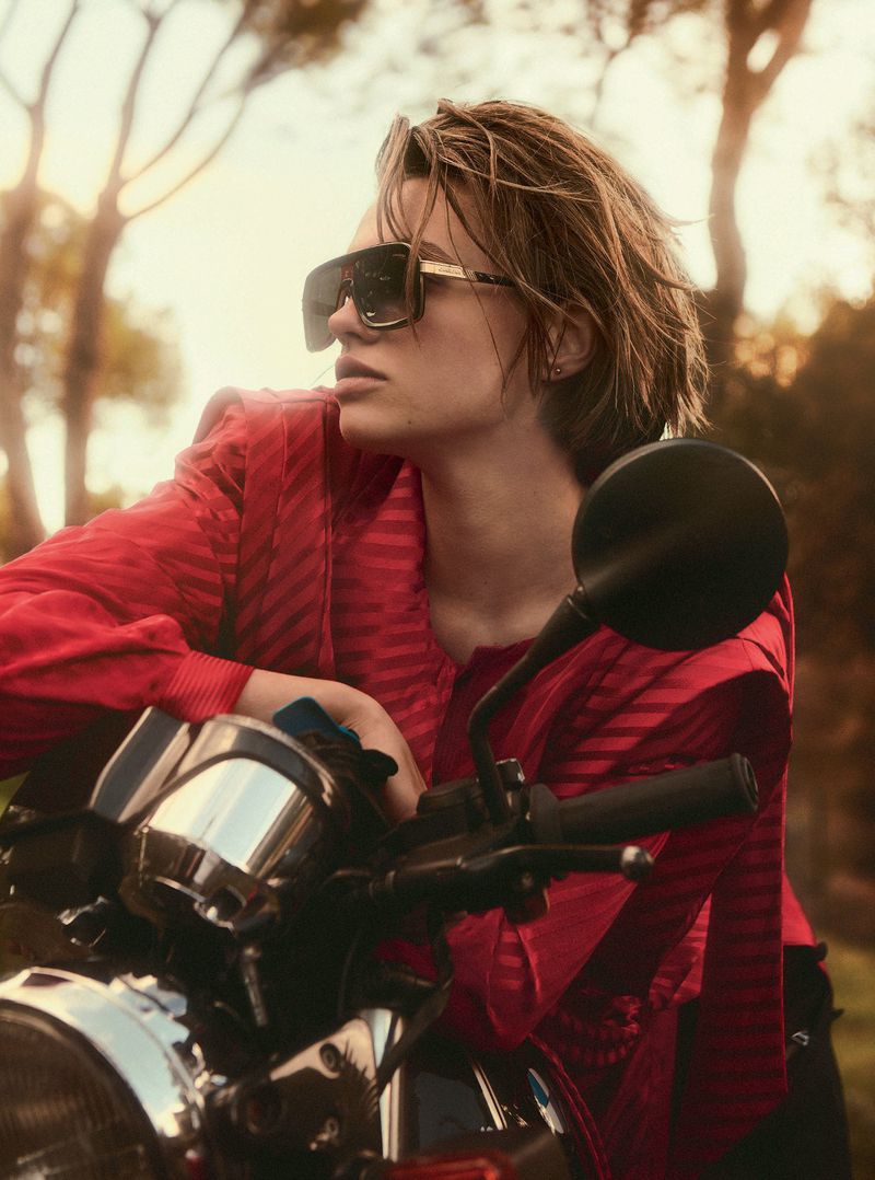 Woman in sunglasses and red jacket leans on a motorcycle in a sunlit outdoor setting with trees, creating a warm, editorial style image.