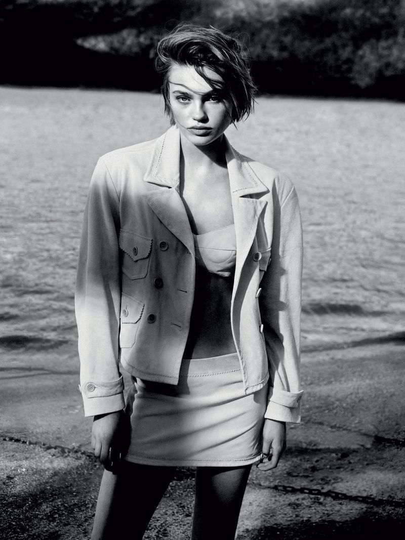 A monochrome portrait of a young woman in a jacket and skirt, standing outdoors with a blurred background of earth and vegetation.