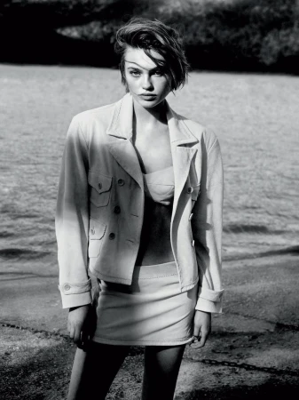 A monochrome portrait of a young woman in a jacket and skirt, standing outdoors with a blurred background of earth and vegetation.
