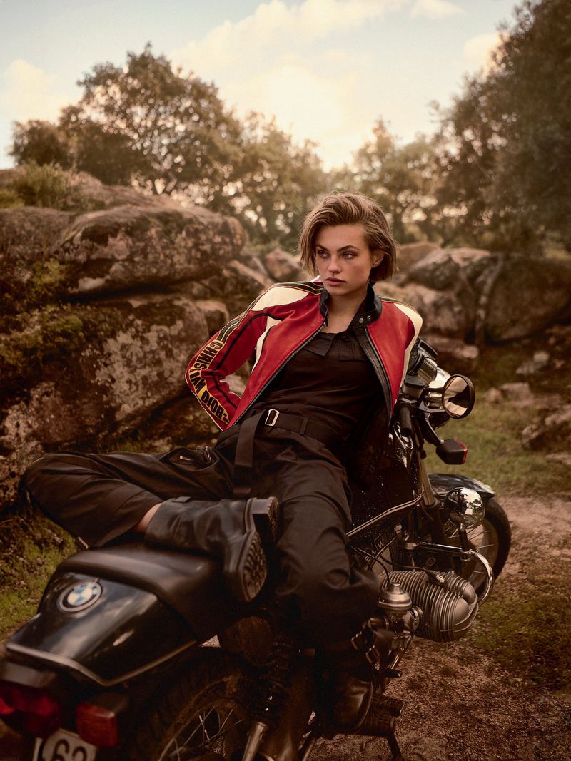 Woman in red Diesel jacket poses on a black BMW motorcycle on a rocky, grassy hillside with trees and a cloudy sky in the background.