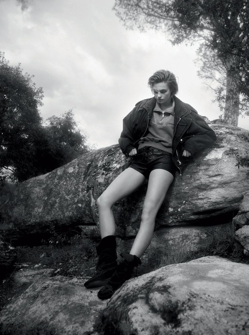Monochrome portrait of a female model wearing black leather shorts, boots, and a jacket, leaning on a rock in a natural setting.
