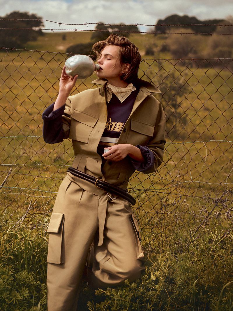 Woman drinking from a canteen in a field, wearing a beige jacket and wide-leg pants, standing near a barbed-wire fence.