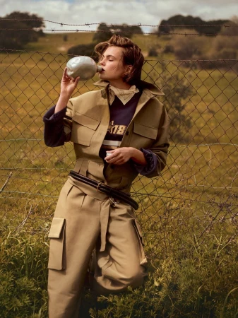 Woman drinking from a canteen in a field, wearing a beige jacket and wide-leg pants, standing near a barbed-wire fence.