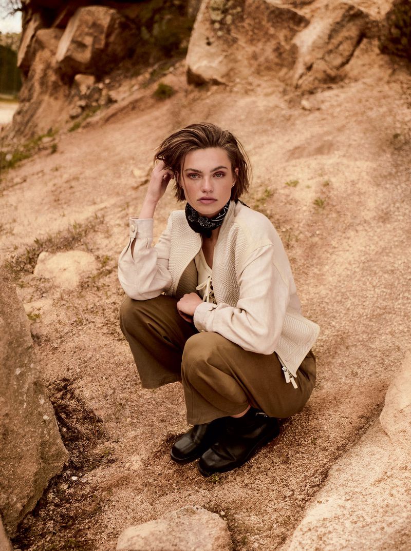 Attractive woman in earth-tone jacket and trousers squats on a rocky surface outdoors, with a desert-like background, posing for fashion shot.