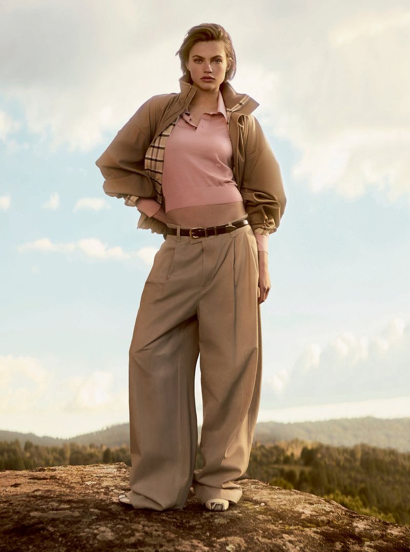 Woman poses on a rocky hill wearing a polo shirt, jacket, and wide-leg pants, with a light blue sky and distant trees in the background.