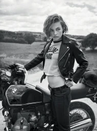 Model Anne Boomgaart wearing a black leather jacket and motorcycle helmet standing by a motorcycle in an outdoor landscape.