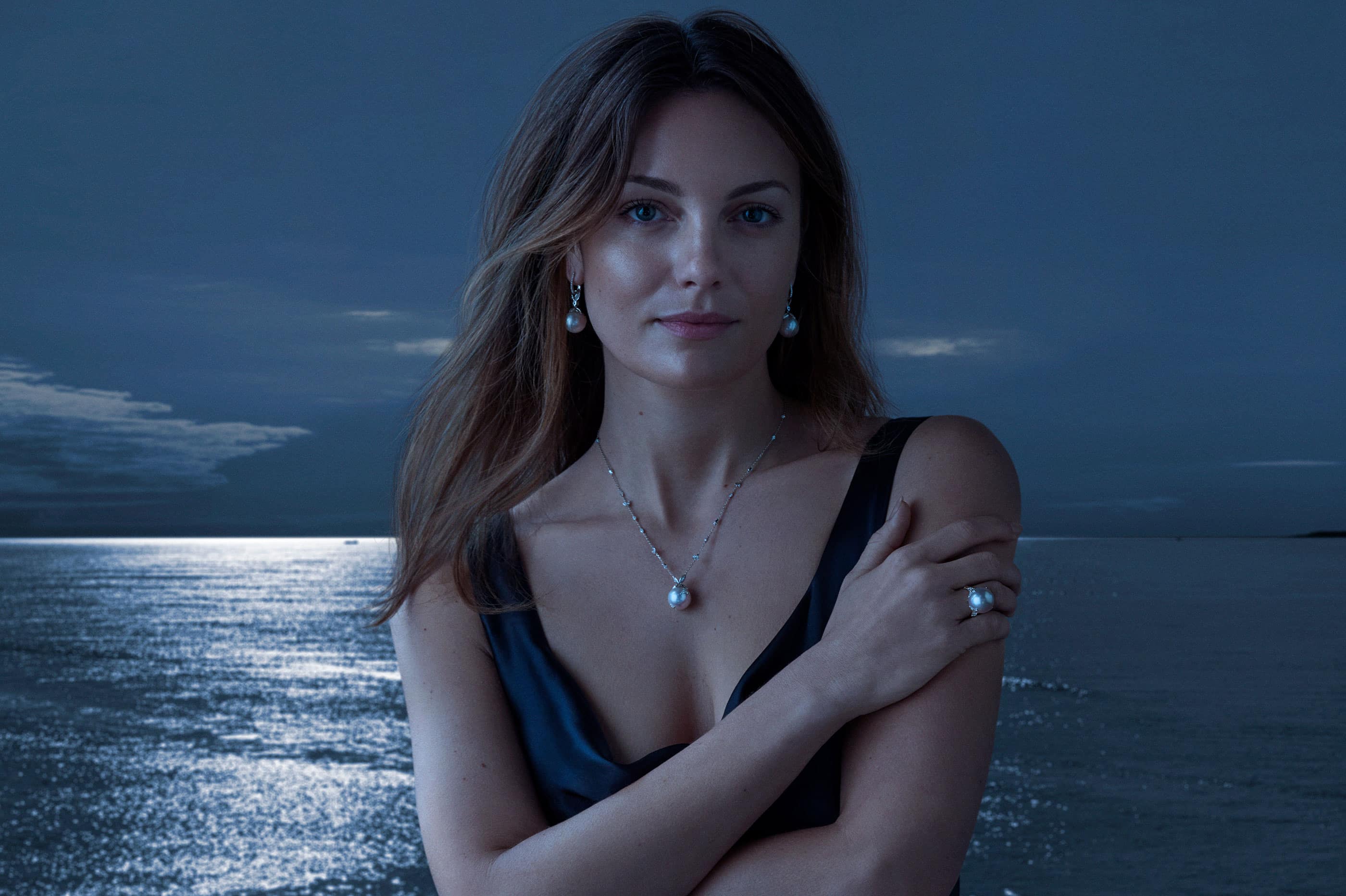 Woman in a black dress wearing pearl earrings, a pearl necklace, and a pearl ring, posing against a dark seascape background.