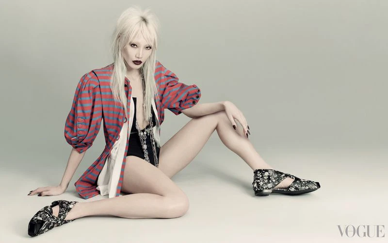 Fashion portrait of a female model with platinum blonde hair, wearing a striped top, black bodysuit, and floral-patterned shoes in a studio.