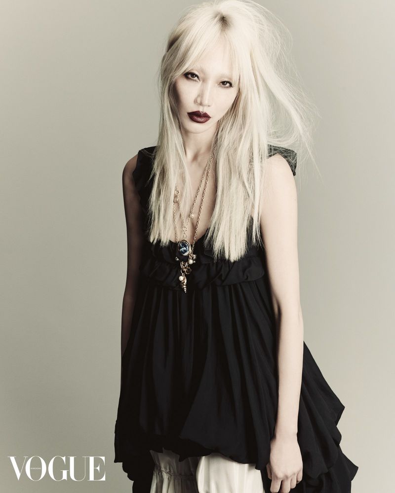 Asian adult female poses in a studio wearing a black top, jewelry, and long white-blonde hair for a Vogue fashion shot.