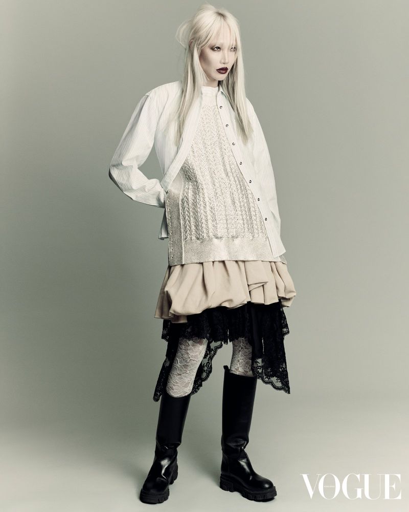 Editorial fashion shot of a woman with platinum blonde hair wearing a white button-down shirt, layered beige and black skirt, and black boots.