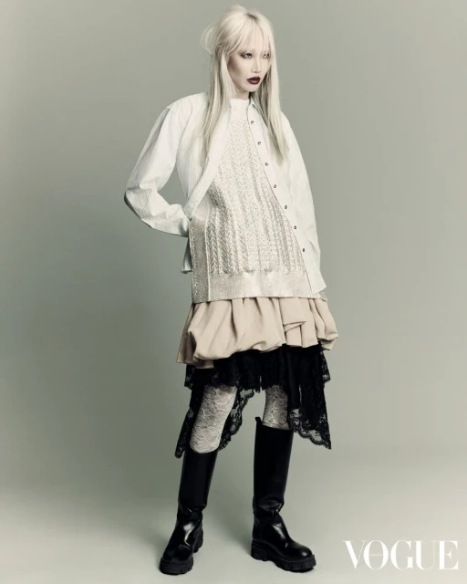 Editorial fashion shot of a woman with platinum blonde hair wearing a white button-down shirt, layered beige and black skirt, and black boots.