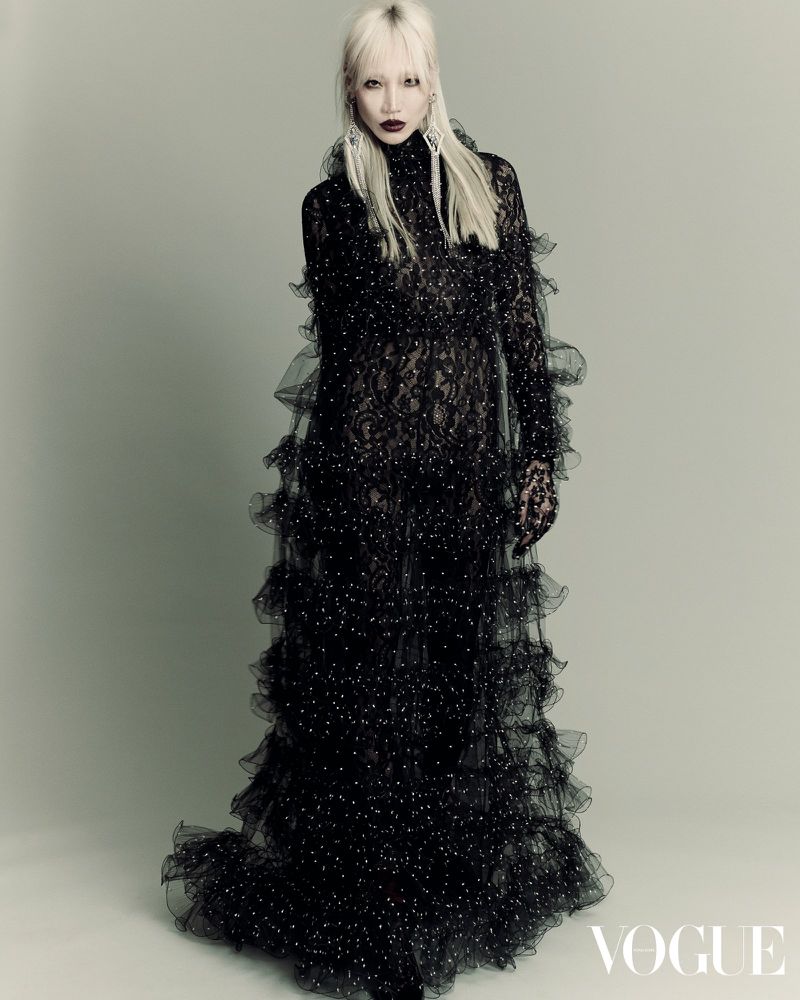 Editorial fashion portrait of an adult female with platinum blonde hair wearing a ruffled black lace gown in a photo studio.