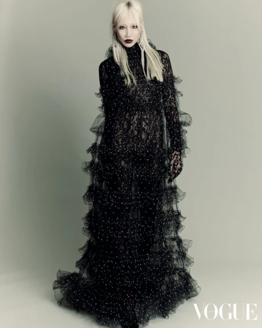 Editorial fashion portrait of an adult female with platinum blonde hair wearing a ruffled black lace gown in a photo studio.
