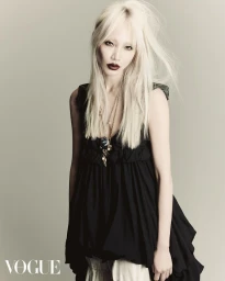 Blonde woman wearing a black ruffled sleeveless dress with layered pendant necklaces against a neutral grey studio background.
