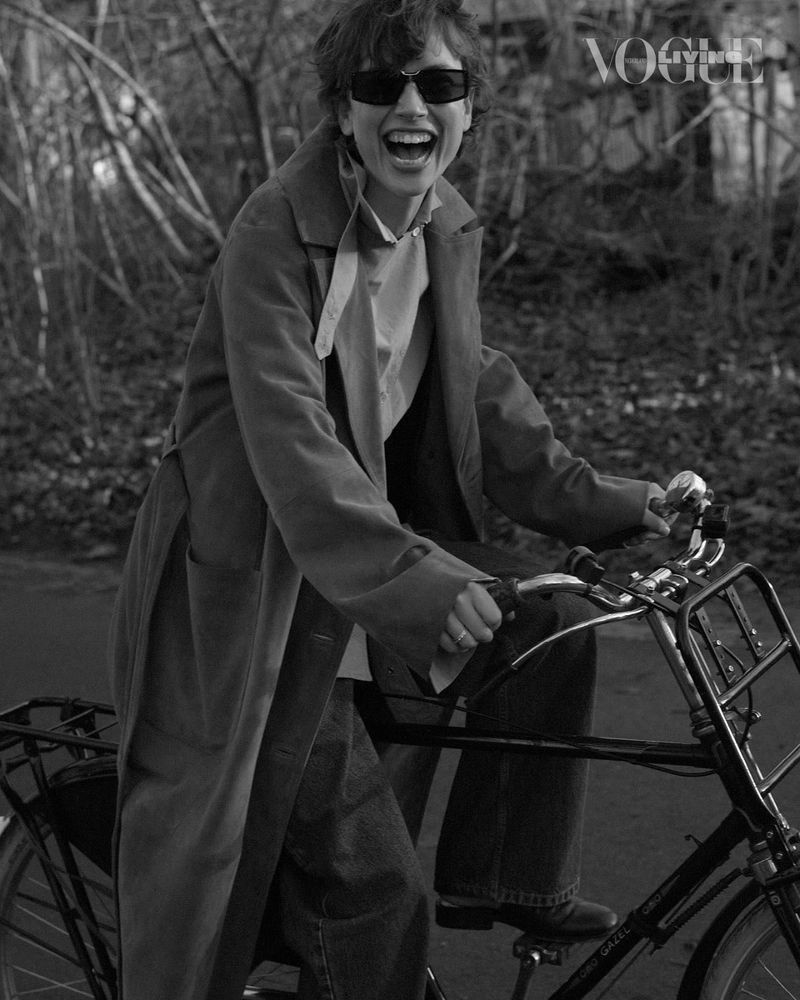A happy woman rides a bicycle outdoors in a black-and-white editorial shot, wearing sunglasses and a long coat, with 'Vogue' logo visible.