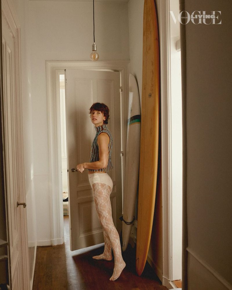 Young adult woman in striped top and lace tights standing indoors near surfboards, styled for a Vogue editorial fashion shoot.